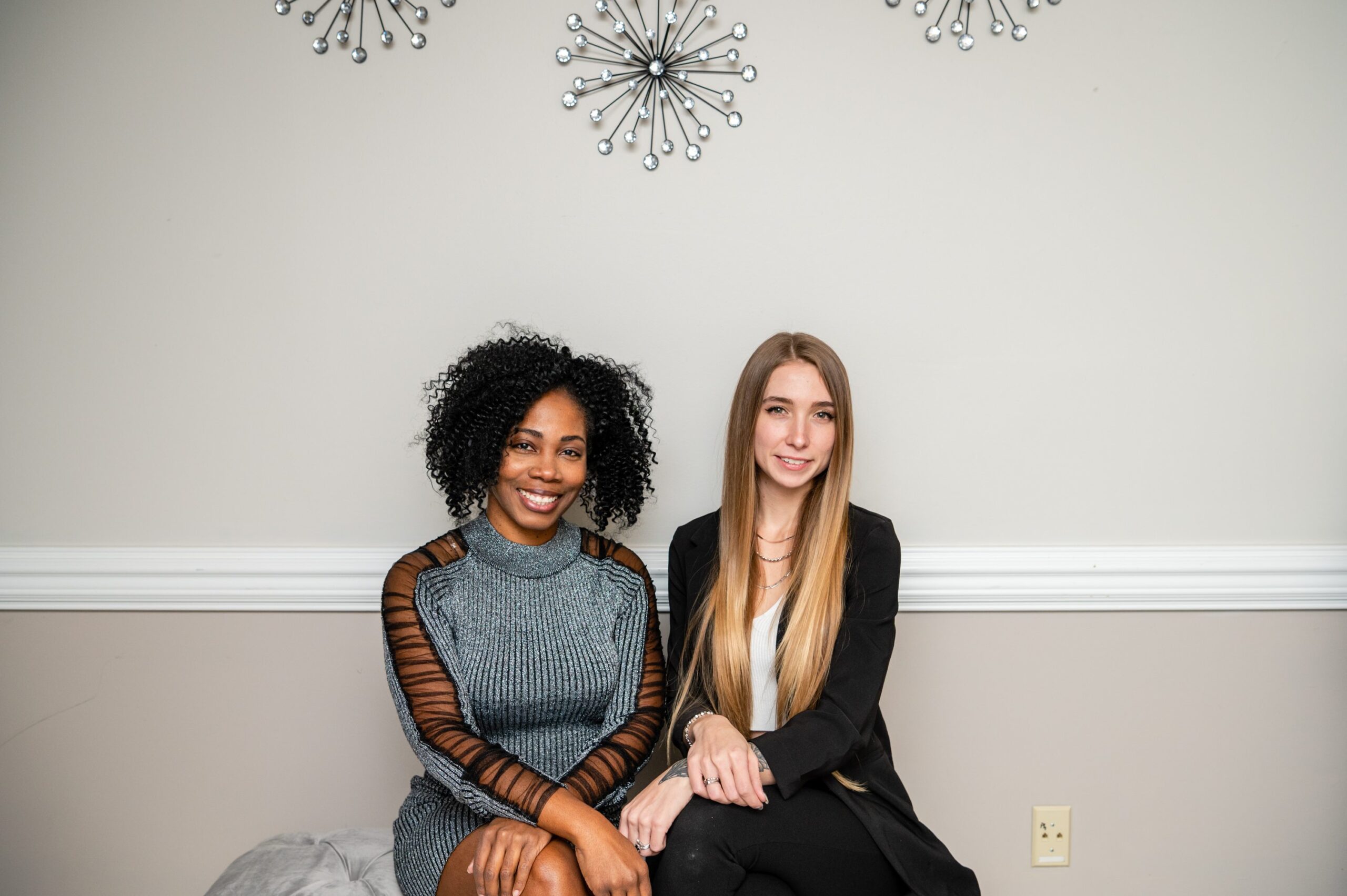 Two women sitting on a bench in front of wall.